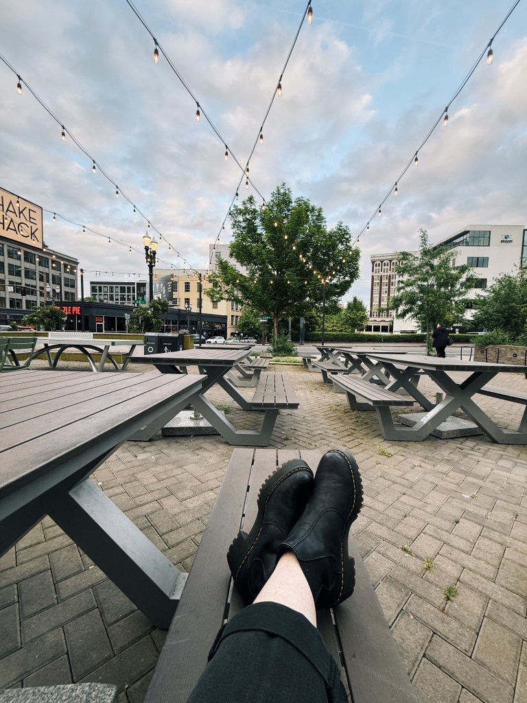 Black boots crossed and resting on a wooden picnic bench under string lights at an outdoor Shake Shack patio in Portland