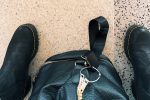 Looking down at black boots and a black leather duffel bag on a speckled terrazzo train station floor