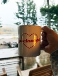 A white ceramic mug with the word "Packwood" inside a heart, sitting in focus with a blurry river, autumn trees, and grey sky in the background.