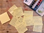 Handwritten post-it notes scattered on a paint-stained wooden desk next to a watercolor palette