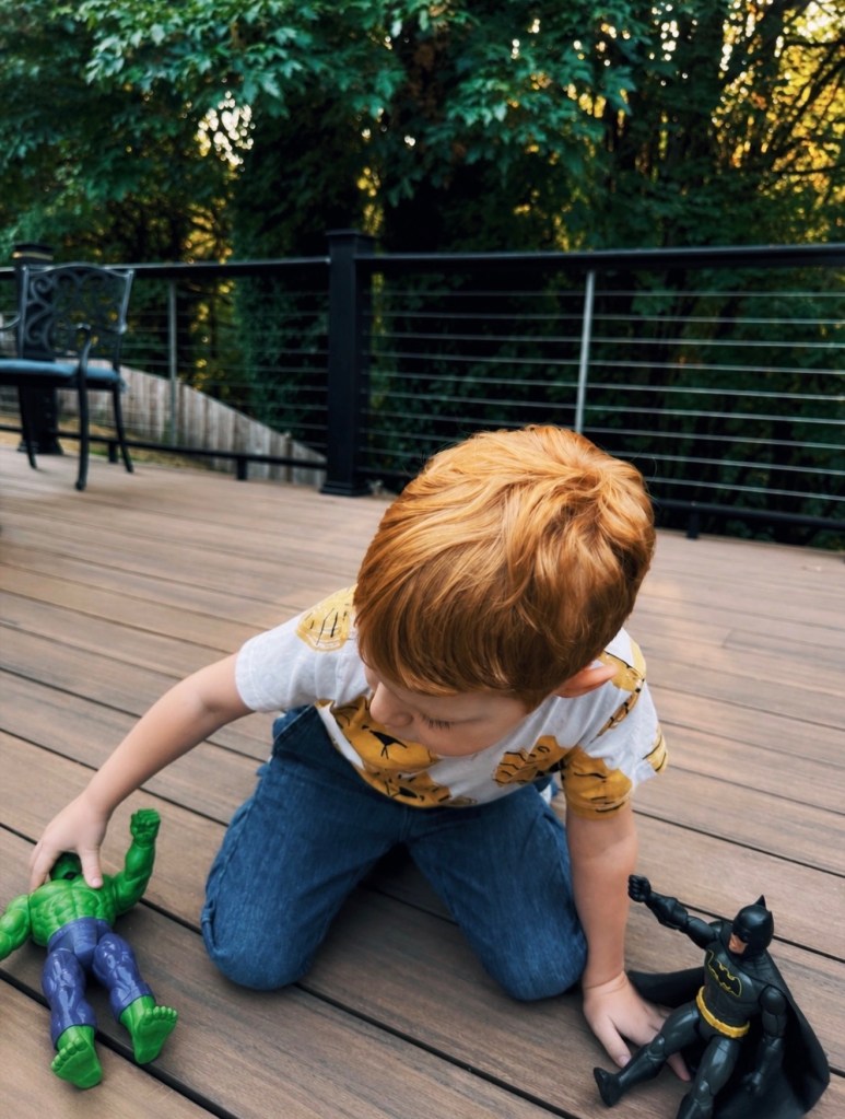 A child playing with foot-long Hulk and Batman figurines on a wooden deck at dusk, with a greenbelt in the background