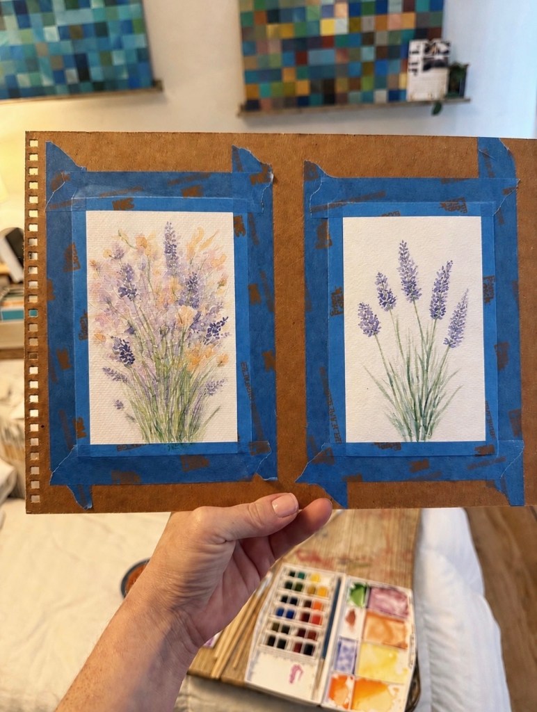 A hand holding a sketchbook with two watercolor paintings of flowers in progress, taped to the page, with a paint palette and mosaic tile background
