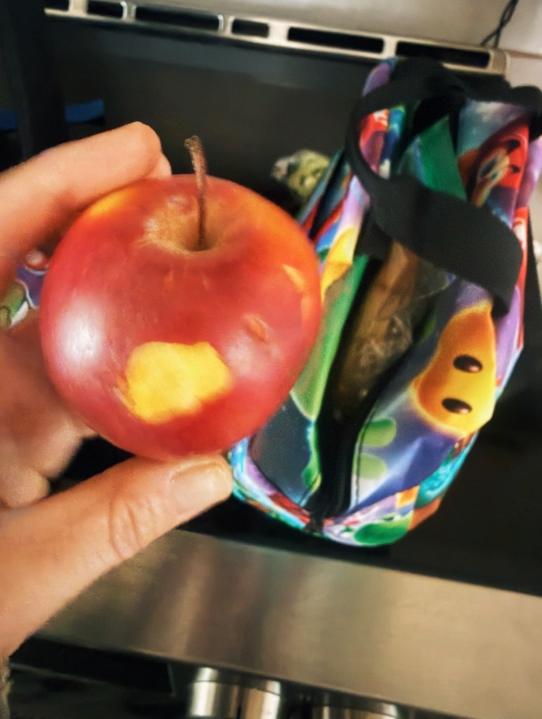A red apple with one tiny bite missing, held in front of a colorful children’s lunch bag.