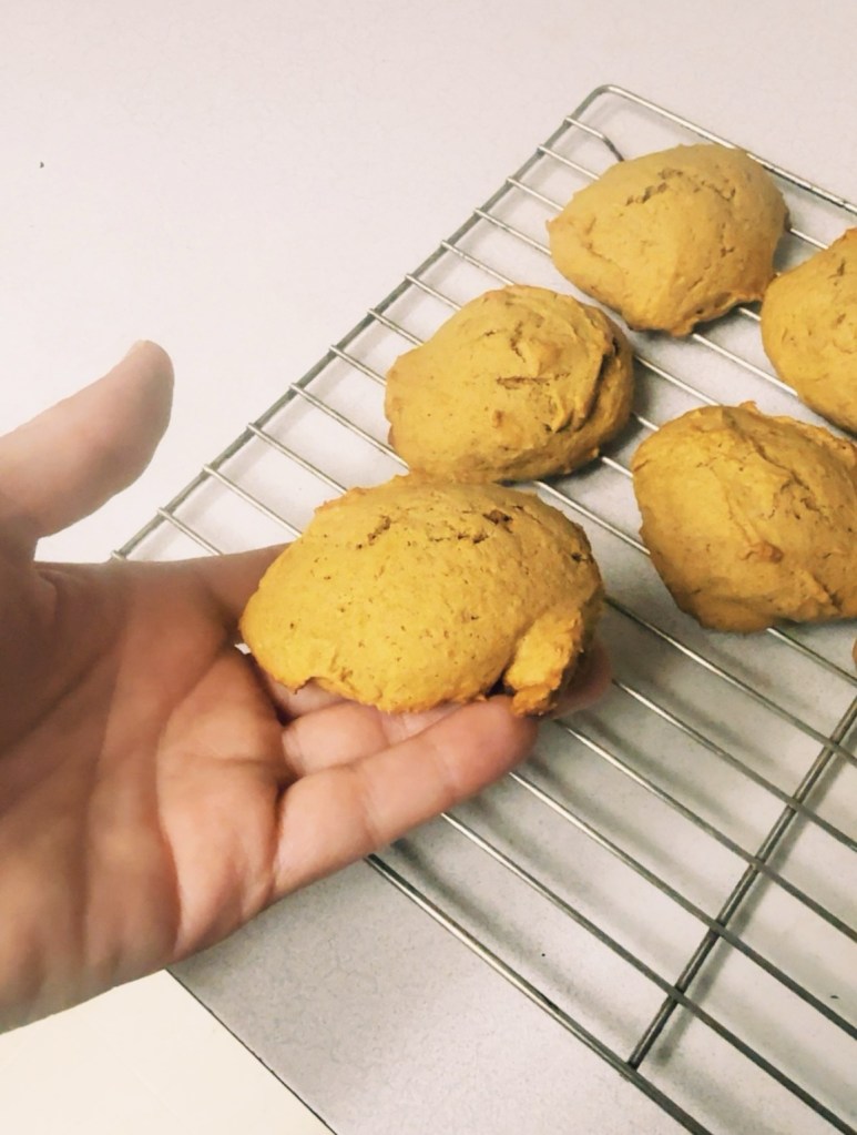 Homemade pumpkin cookies cooling on a wire rack, with one thick, round cookie held in a hand.